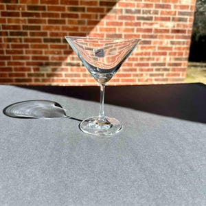 May include: A clear glass martini glass with a stem and a wide bowl. The glass is sitting on a gray surface.