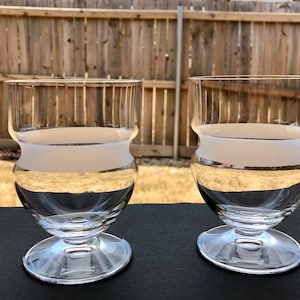 May include: Two clear glass drinking glasses with a frosted white band around the middle. The glasses have a wide base and a rounded bowl.
