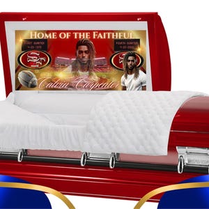 May include: A red casket with a white quilted lining. The lid is open and features a custom design with the words "Home of the Faithful" and a photo of a football player. The casket is decorated with silver accents.