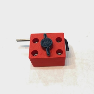 May include: A red 3D-printed object with a square base and a black handle. The object has a central black knob and four circular holes. A silver metal rod extends from one side. The object is on a white surface.
