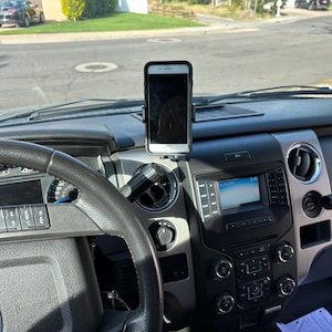 May include: A black and silver car phone mount with a smartphone attached, positioned on a vehicle's dashboard. The interior features a steering wheel, dashboard controls, and a digital display. The background shows a residential street.