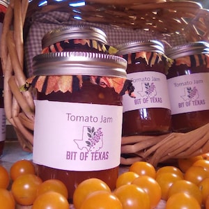 May include: Several jars of tomato jam labeled "Bit of Texas" are arranged in a woven basket and on a surface, alongside a pile of small, yellow tomatoes. The jars have decorative fabric tops.