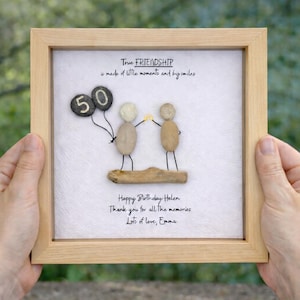 May include: Framed artwork with a light wood frame. The artwork features pebble art of two figures holding hands, with a '50' balloon. Text reads 'True Friendship' and a birthday message.