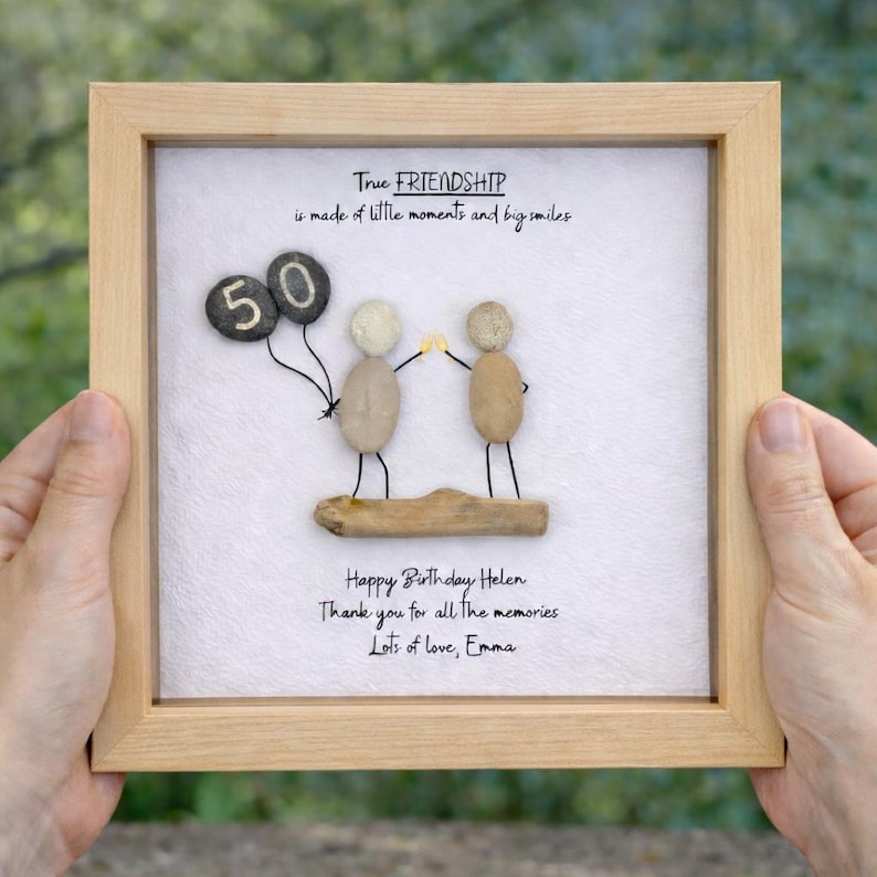 May include: Framed artwork with a light wood frame. The artwork features pebble art of two figures holding hands, with a '50' balloon. Text reads 'True Friendship' and a birthday message.