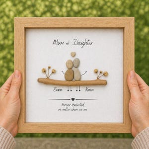 Puede incluir: Una obra de arte enmarcada con un marco de madera clara. La obra presenta una rama con dos figuras de guijarros, un coraz&oacute;n y el texto "Mum + Daughter". Los nombres "Emma" y "Karen" est&aacute;n debajo, con el texto "Forever connected no matter where we are".