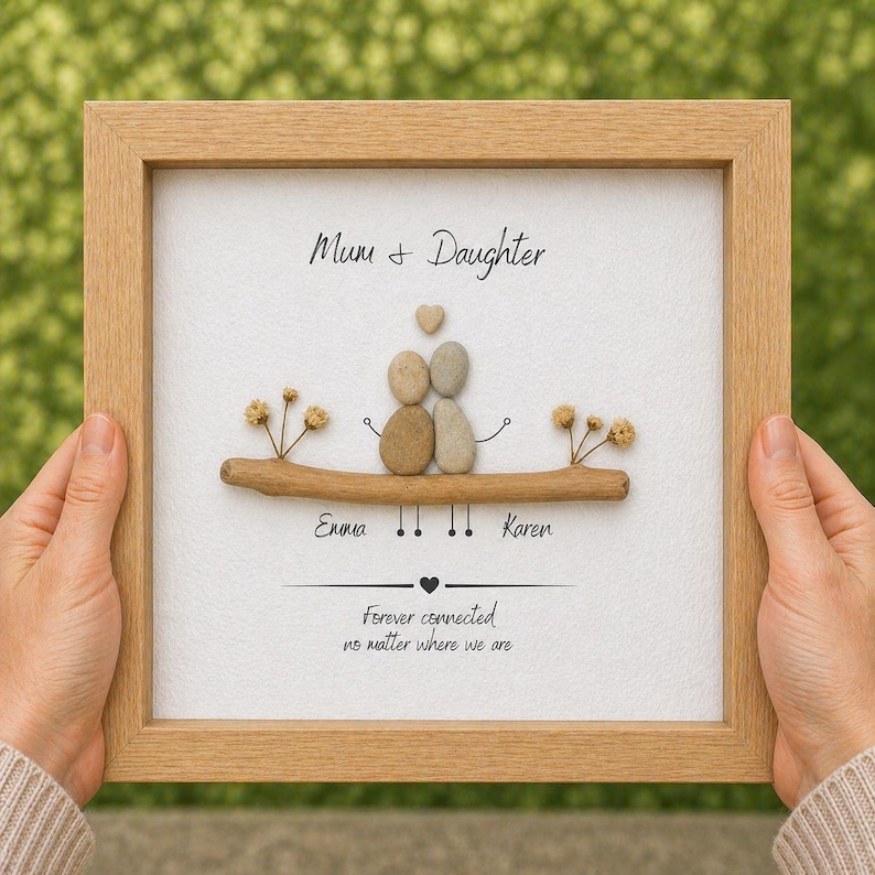 Puede incluir: Una obra de arte enmarcada con un marco de madera clara. La obra presenta una rama con dos figuras de guijarros, un coraz&oacute;n y el texto "Mum + Daughter". Los nombres "Emma" y "Karen" est&aacute;n debajo, con el texto "Forever connected no matter where we are".