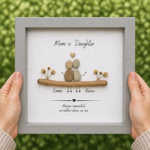 Puede incluir: Una obra de arte enmarcada con un borde gris presenta un dise&ntilde;o de arte de guijarros. El arte representa dos figuras hechas de guijarros, un coraz&oacute;n y las palabras "Mum + Daughter". Los nombres "Emma" y "Karen" tambi&eacute;n est&aacute;n incluidos.