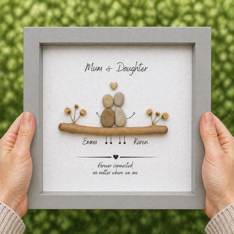 Puede incluir: Una obra de arte enmarcada con un borde gris presenta un dise&ntilde;o de arte de guijarros. El arte representa dos figuras hechas de guijarros, un coraz&oacute;n y las palabras "Mum + Daughter". Los nombres "Emma" y "Karen" tambi&eacute;n est&aacute;n incluidos.