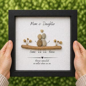 Puede incluir: Una obra de arte enmarcada con un marco negro, que presenta un dise&ntilde;o de arte de guijarros. El arte representa a una madre y una hija con un coraz&oacute;n encima de ellas. El texto dice "Mum + Daughter" y "Forever connected no matter where we are". Tambi&eacute;n se incluyen los nombres "Emma" y "Karen".