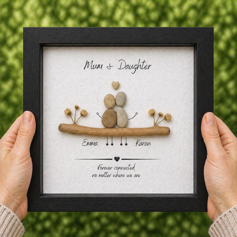 Puede incluir: Una obra de arte enmarcada con un marco negro, que presenta un dise&ntilde;o de arte de guijarros. El arte representa a una madre y una hija con un coraz&oacute;n encima de ellas. El texto dice "Mum + Daughter" y "Forever connected no matter where we are". Tambi&eacute;n se incluyen los nombres "Emma" y "Karen".