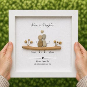 Puede incluir: Una obra de arte enmarcada en blanco. Presenta dos figuras de guijarros sobre una rama, con los nombres "Emma" y "Karen" debajo. El texto dice "Mum & Daughter" y "Forever connected no matter where we are".