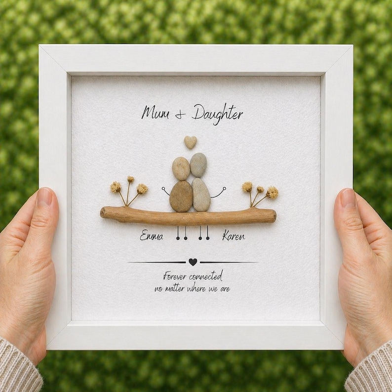 Puede incluir: Una obra de arte enmarcada en blanco. Presenta dos figuras de guijarros sobre una rama, con los nombres "Emma" y "Karen" debajo. El texto dice "Mum & Daughter" y "Forever connected no matter where we are".