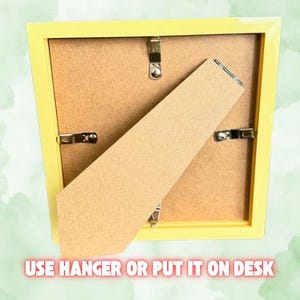 Puede incluir: Parte trasera de un marco de fotos amarillo con respaldo de cart&oacute;n y clips met&aacute;licos. El marco tiene un caballete y un colgador. El texto en la parte inferior dice "USE HANGER OR PUT IT ON DESK".