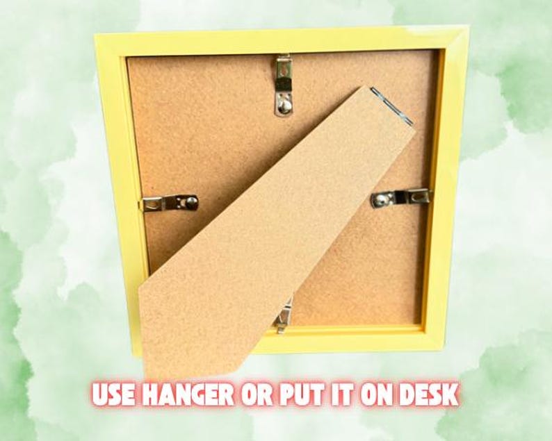 Puede incluir: Parte trasera de un marco de fotos amarillo con respaldo de cart&oacute;n y clips met&aacute;licos. El marco tiene un caballete y un colgador. El texto en la parte inferior dice "USE HANGER OR PUT IT ON DESK".