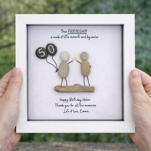May include: A framed artwork with a white border. The artwork features pebble art of two figures holding hands, with a '50' balloon. Text reads 'True FRIENDSHIP', 'Happy Birthday Helen', and a personal message. The background is white.