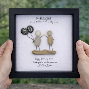 May include: A framed artwork with a black frame. The artwork features pebble art of two figures holding hands, with a '50' balloon. Text reads: 'True FRIENDSHIP is made of little moments and big smiles.' and a birthday message.