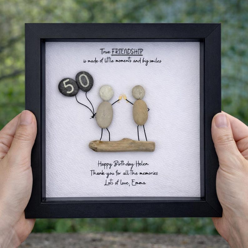 May include: A framed artwork with a black frame. The artwork features pebble art of two figures holding hands, with a '50' balloon. Text reads: 'True FRIENDSHIP is made of little moments and big smiles.' and a birthday message.
