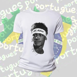 Puede incluir: Camiseta blanca con un retrato en blanco y negro de una persona que lleva una cinta en la cabeza con el texto "100% JESUS". La camiseta tiene cuello redondo y mangas cortas. El fondo incluye la bandera brasileña borrosa y la palabra "Portugués".
