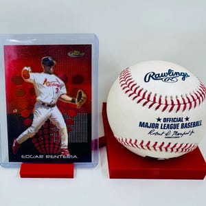 May include: A baseball card featuring Edgar Renteria in a Cardinals uniform, displayed in a clear holder. Beside it, a Rawlings Major League Baseball rests on a red stand. The baseball has red stitching and official markings.