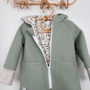 Könnte beinhalten: Salbeigrüner Kapuzenjacke mit weißem Reißverschluss und zwei Vordertaschen. Die Jacke hat ein floral gemustertes Futter und hellgraue Bündchen. Das Etikett "jomisa" ist sichtbar. Die Jacke hängt an einem Holzbügel.