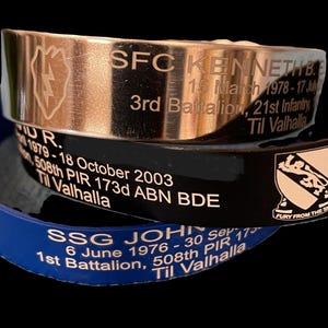 May include: Three stacked bracelets with engraved text and a military insignia. The top bracelet is silver with a gold inscription, the middle bracelet is black with a gold inscription, and the bottom bracelet is blue with a gold inscription. The text on the bracelets reads: "SFC KENNETH B. 15 March 1978-17 July 3rd Battalion, 21st Infantry Til Valhalla", "ID R. 1979-18 October 2003 508th PIR 173d ABN BDE Til Valhalla", and "SSG JOHN 6 June 1976-30 Sep 1979 1st Battalion, 508th PIR Til Valhalla". The military insignia is a gold shield with a lion and the text "Fury From The Sky".