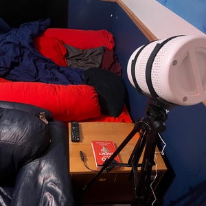 May include: A white cylindrical light with black straps is mounted on a black tripod. A red book with the title "The Enemy" is on a wooden nightstand. A remote control is also on the nightstand. A bed with red and blue bedding is in the background.