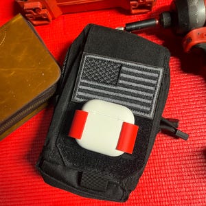 May include: Black tactical pouch with a gray and black American flag patch. A white AirPod case is secured with red straps. The pouch is on a red surface, with tools and a brown wallet in the background.