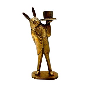 May include: A brass-colored figurine of a rabbit dressed in a suit, holding a small top hat. The rabbit has long ears and is standing on a rectangular base. The figurine is likely a decorative item or a candle holder.