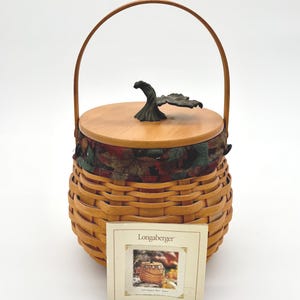 Peut inclure: Un panier en osier avec un couvercle et une poignée en bois. Le couvercle est surmonté d'un élément décoratif en forme de feuille. Le panier a une doublure en tissu avec un motif sur le thème de l'automne. Une petite carte avec le logo Longaberger est devant le panier.