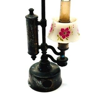 May include: A miniature antique-style lamp with a black metal base and frame. The lamp features a small, white lampshade adorned with pink floral patterns. The lamp has a cylindrical glass chimney and a decorative top.
