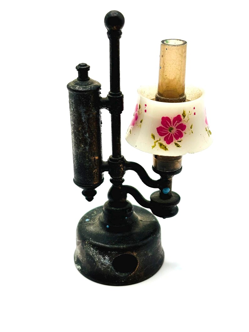 May include: A miniature antique-style lamp with a black metal base and frame. The lamp features a small, white lampshade adorned with pink floral patterns. The lamp has a cylindrical glass chimney and a decorative top.