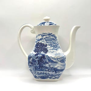 May include: A white and blue ceramic coffee pot with a lid. The pot features a scenic blue and white landscape design with a tree, bridge, and water. The handle and spout are white.