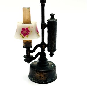 May include: A miniature antique lamp with a white lampshade adorned with pink floral designs. The lamp has a dark bronze finish and a cylindrical reservoir. The lamp is a decorative item.
