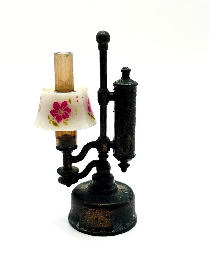 May include: A miniature antique lamp with a white lampshade adorned with pink floral designs. The lamp has a dark bronze finish and a cylindrical reservoir. The lamp is a decorative item.