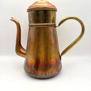 May include: A vintage copper coffee pot with a brass handle and lid. The pot has a long spout and a rounded body. The copper has a warm, aged patina with hints of red and green. The handle is curved and the lid has a small finial.