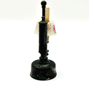 May include: A miniature antique lamp with a dark, aged metal base and stem. The lamp features a small lampshade with a floral pattern in pink and white. The lamp is a decorative item.