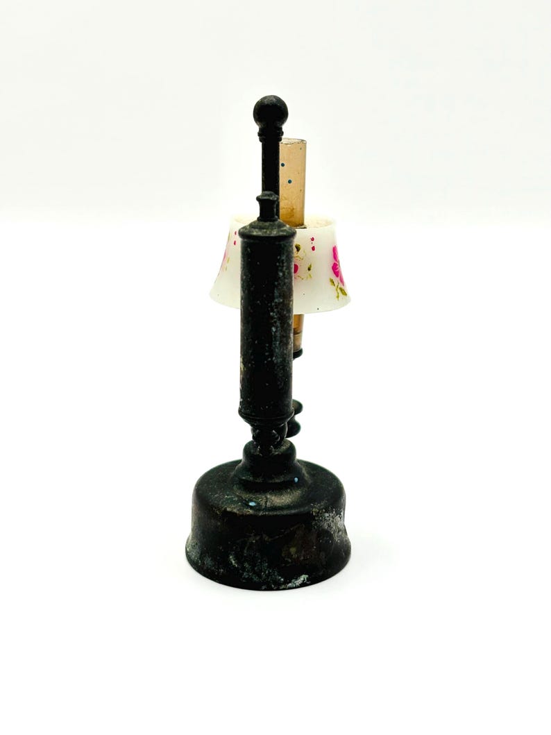 May include: A miniature antique lamp with a dark, aged metal base and stem. The lamp features a small lampshade with a floral pattern in pink and white. The lamp is a decorative item.