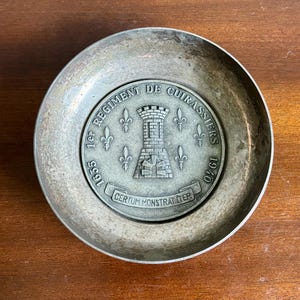 以下が含まれることがあります： 中央にメダリオンが付いた銀色の金属製の皿。メダリオンには城、フルール・ド・リスのシンボル、フランス語のテキスト（「1er REGIMENT DE CUIRASSIERS 1635-1940」および「CERTUM MONSTRAT TIER」を含む）が描かれています。