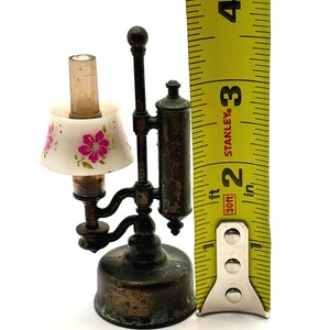 May include: Miniature antique lamp with a white lampshade decorated with pink floral designs. The lamp is made of metal and measures approximately 3 inches tall.