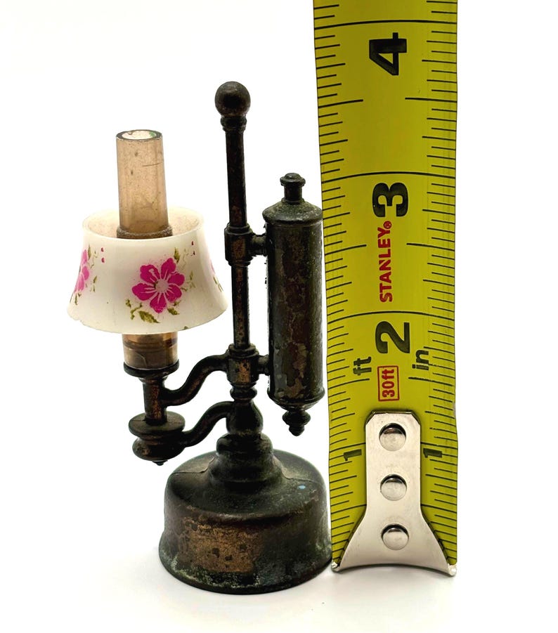 May include: Miniature antique lamp with a white lampshade decorated with pink floral designs. The lamp is made of metal and measures approximately 3 inches tall.