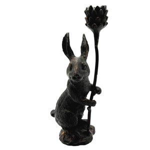 May include: A dark bronze-colored rabbit figurine holding a stylized flower. The rabbit is seated with its ears erect, and the flower is held upright. The figurine has a textured surface and is set against a white background.