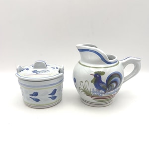 May include: A white ceramic creamer and lidded container set. The creamer features a blue and green rooster design. The container has a matching blue and green leaf pattern. Both pieces have blue and green accents.