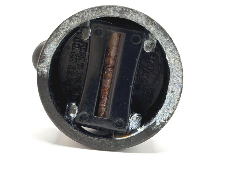 May include: Close-up of a dark metal bottle stopper. The stopper has a circular base with a black plastic insert and a small, rounded handle. The text "MADE IN SPAIN" is visible on the base.