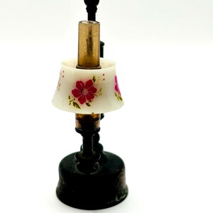 May include: A miniature lamp with a white lampshade adorned with pink floral designs. The lamp has a brass-colored central column and a black base. The lamp is a decorative item.