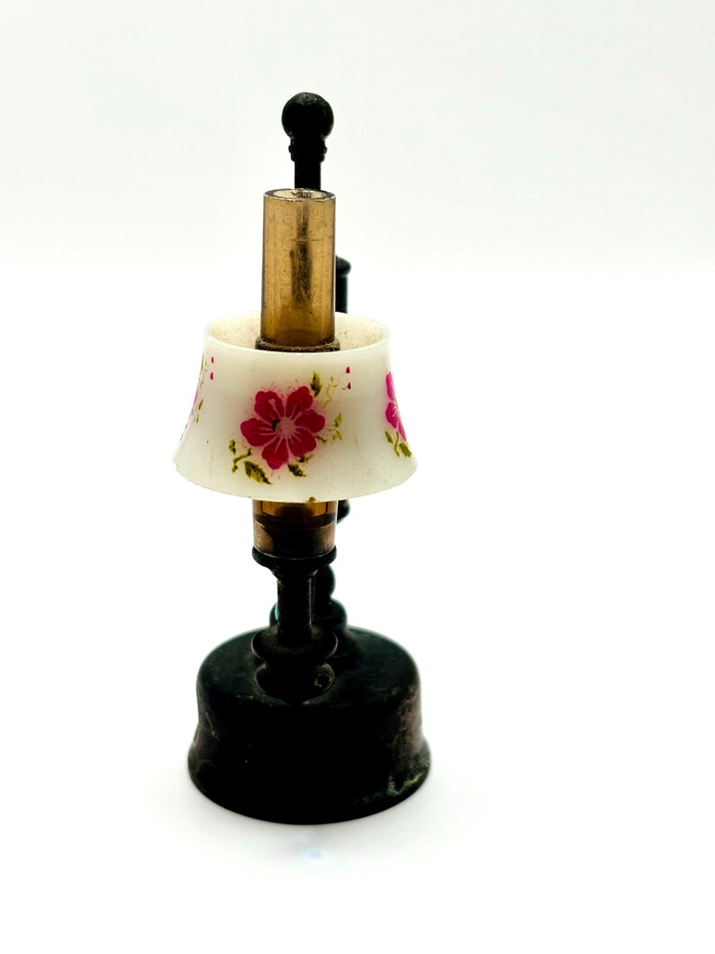 May include: A miniature lamp with a white lampshade adorned with pink floral designs. The lamp has a brass-colored central column and a black base. The lamp is a decorative item.