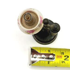 May include: An antique, white ceramic candle holder with pink floral designs. The candle holder is attached to a dark metal base. A yellow measuring tape shows the candle holder is approximately 2 inches in length.