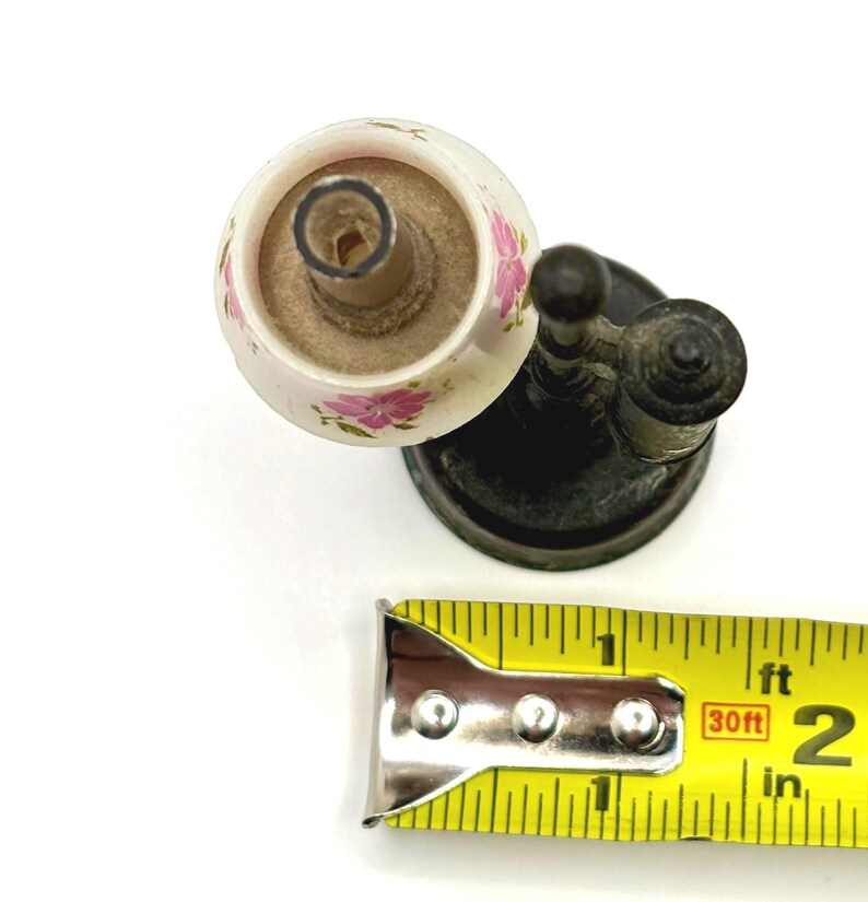 May include: An antique, white ceramic candle holder with pink floral designs. The candle holder is attached to a dark metal base. A yellow measuring tape shows the candle holder is approximately 2 inches in length.