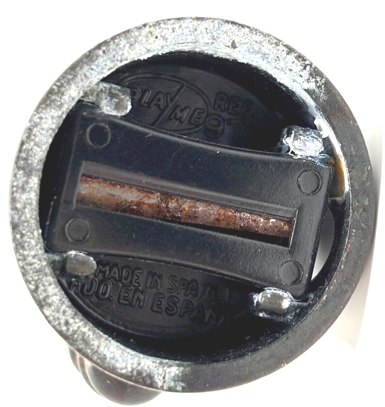 May include: Close-up of a vintage pencil sharpener. The sharpener is black with a rusty blade and is housed in a metallic casing. The text "PLAY ME" and "MADE IN SPAIN" are visible.