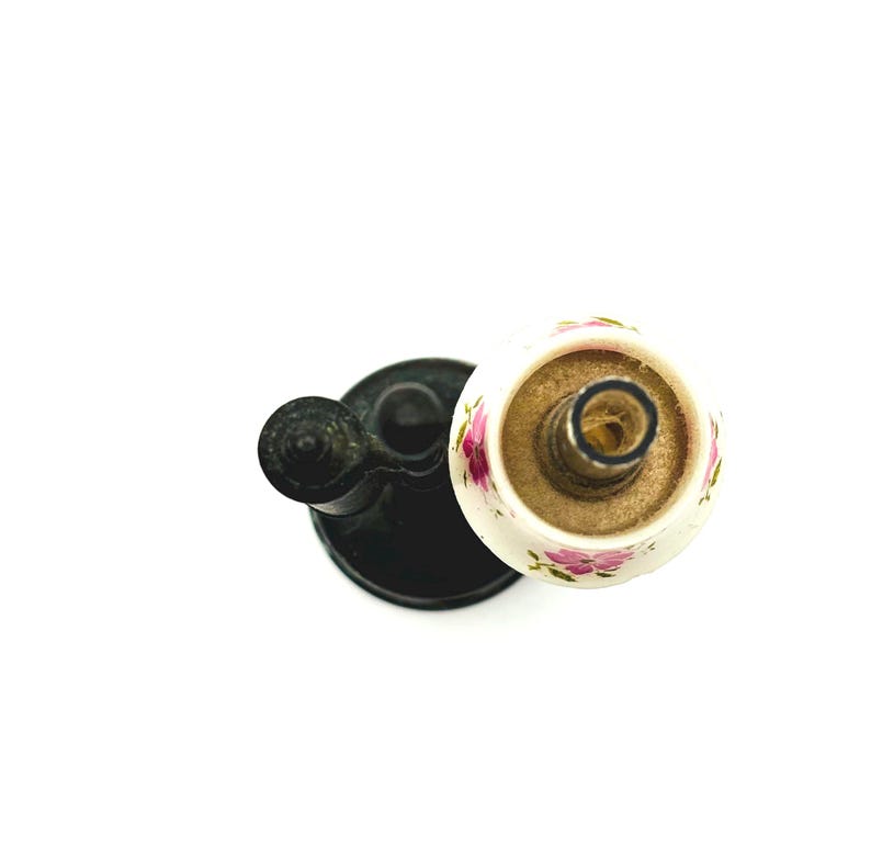 May include: Overhead view of a black metal candle holder with a white and floral-patterned candle cup. The candle cup has pink flowers and green leaves. The candle holder is on a white background.