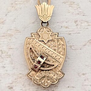 May include: A gold-toned pendant with a Star of David and intricate engraved details. The pendant features a shield shape with a decorative floral design at the bottom and a small band with three red gemstones.